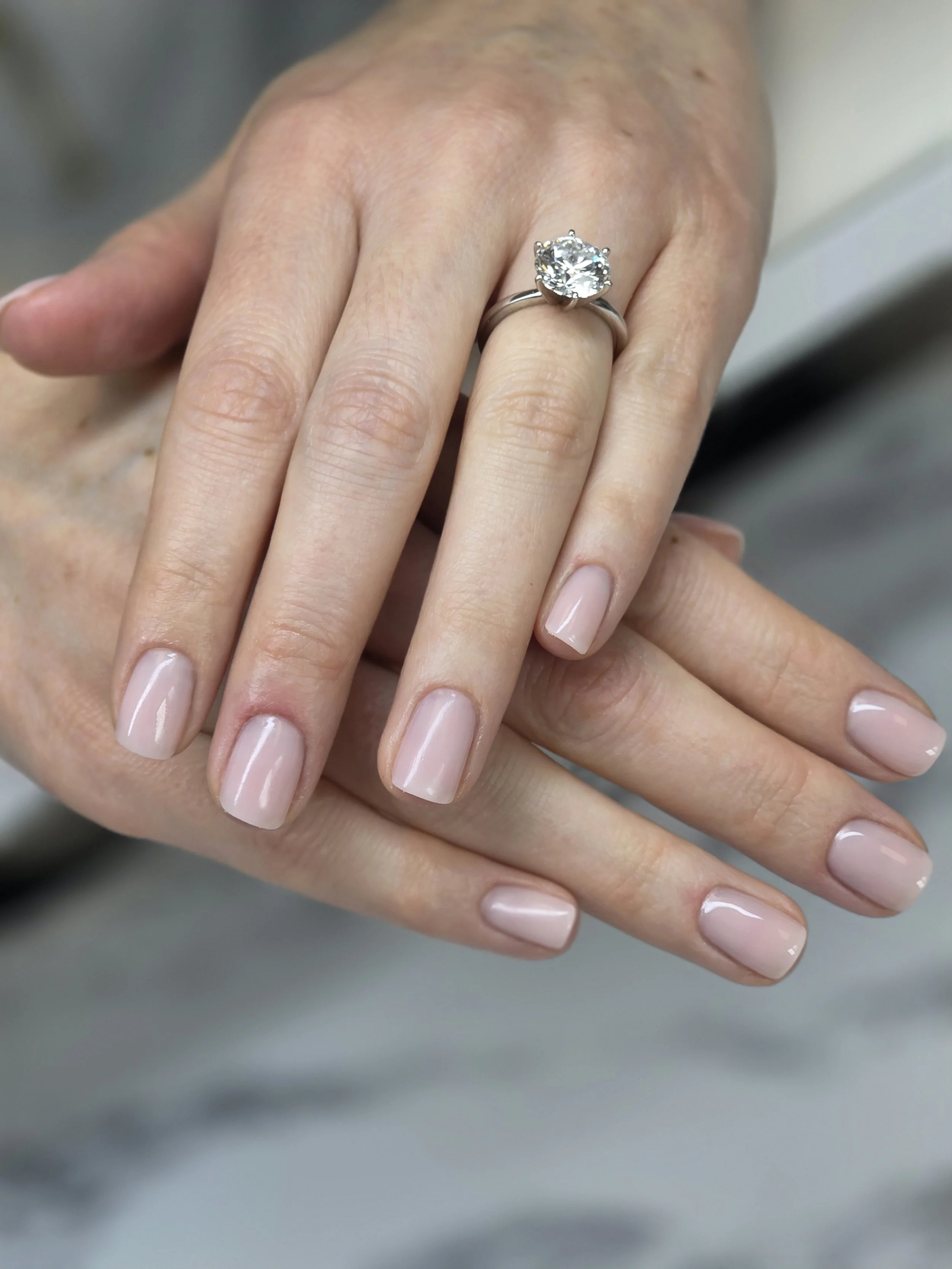 A close-up of a woman's hand wearing a big diamond engagement ring with a silver band. The nails are manicured with light pink polish.