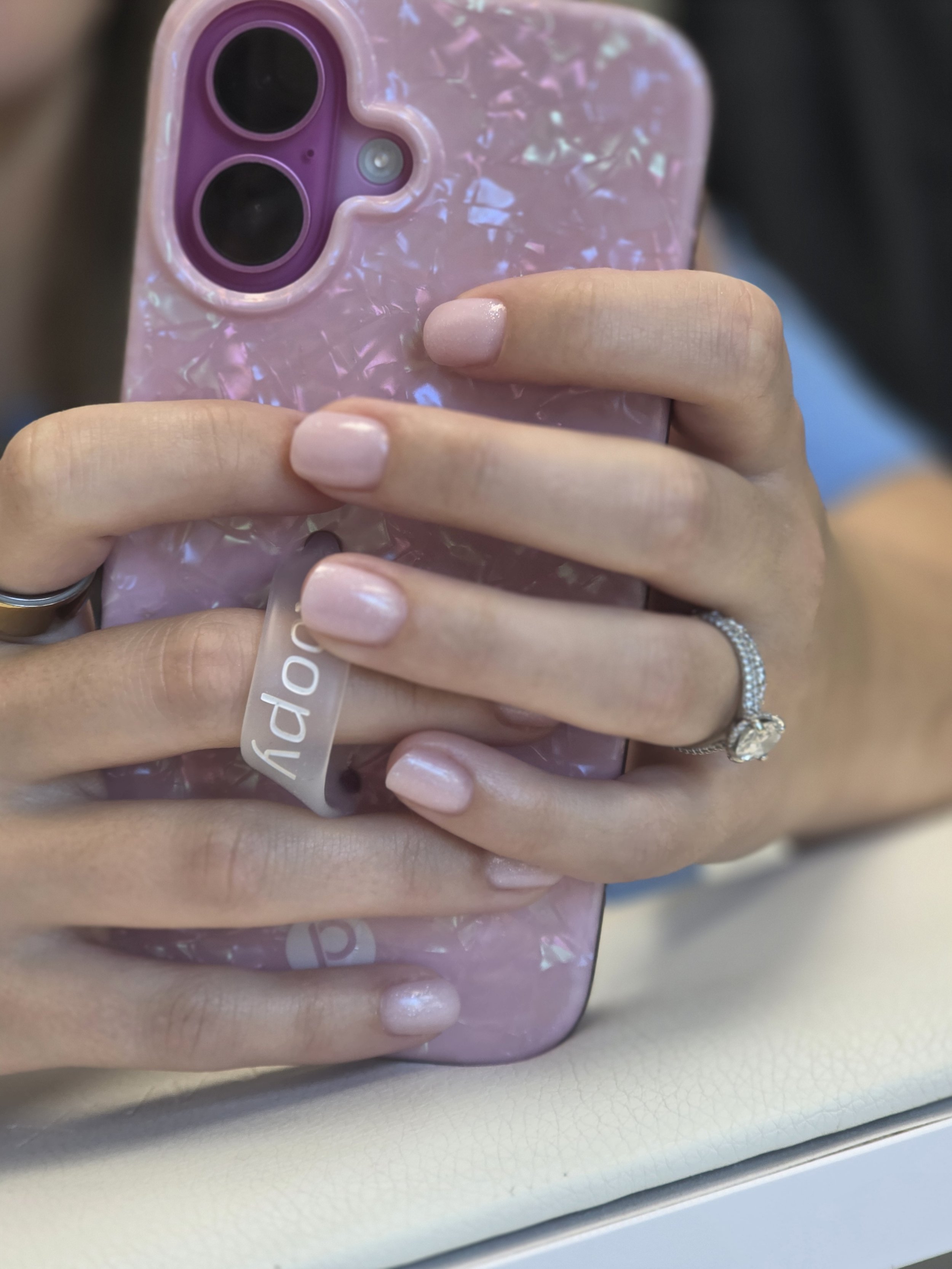Close-up of a person's hand holding a pink iridescent phone with a protective case. The person has well-manicured nails with light pink polish and is wearing a silver ring with a clear gemstone on their finger.