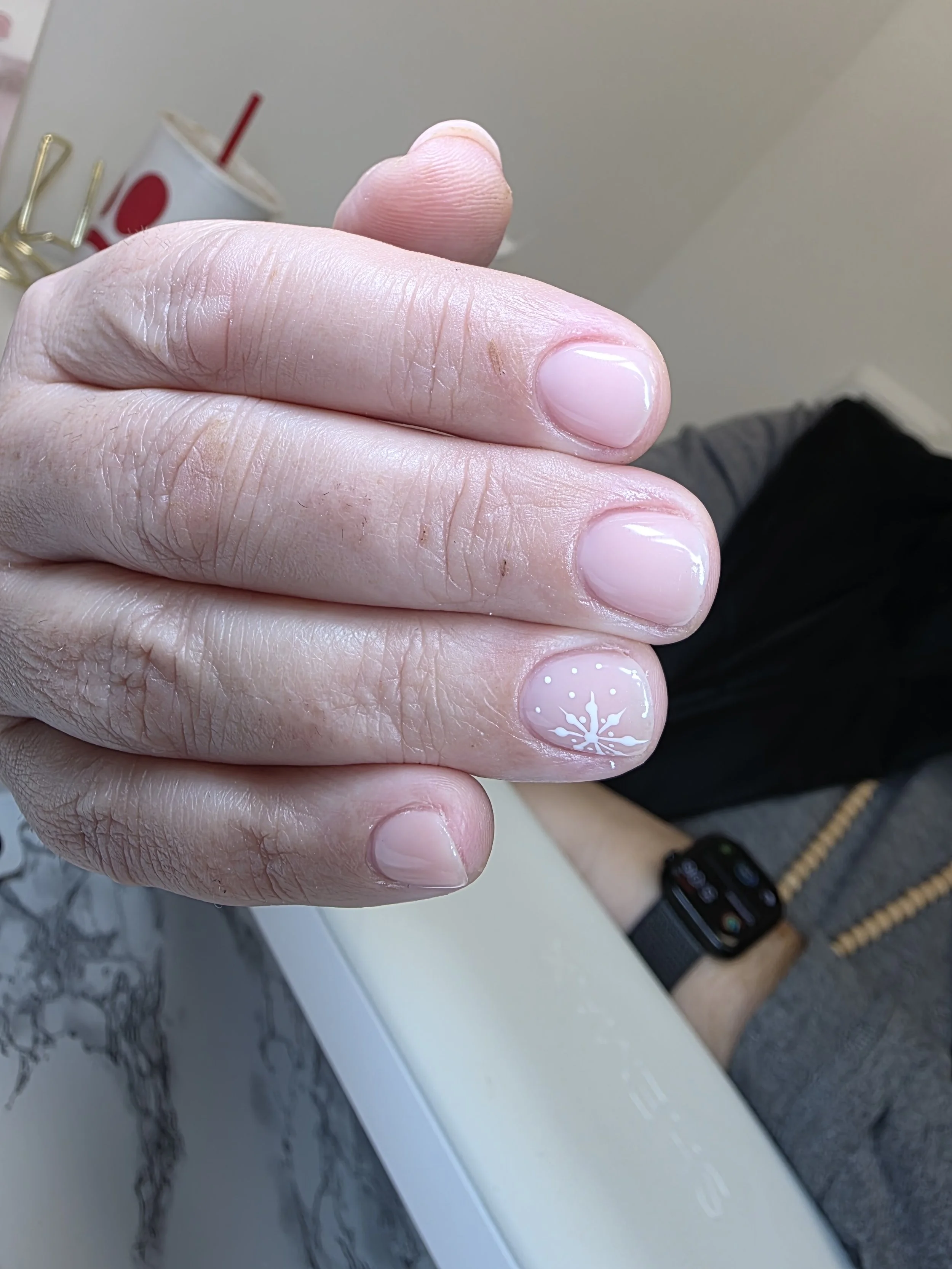 Close-up of a person's hand with neatly manicured nails, one of which has a white snowflake design, resting on a marble countertop. A cup with a straw and a food court in the background.