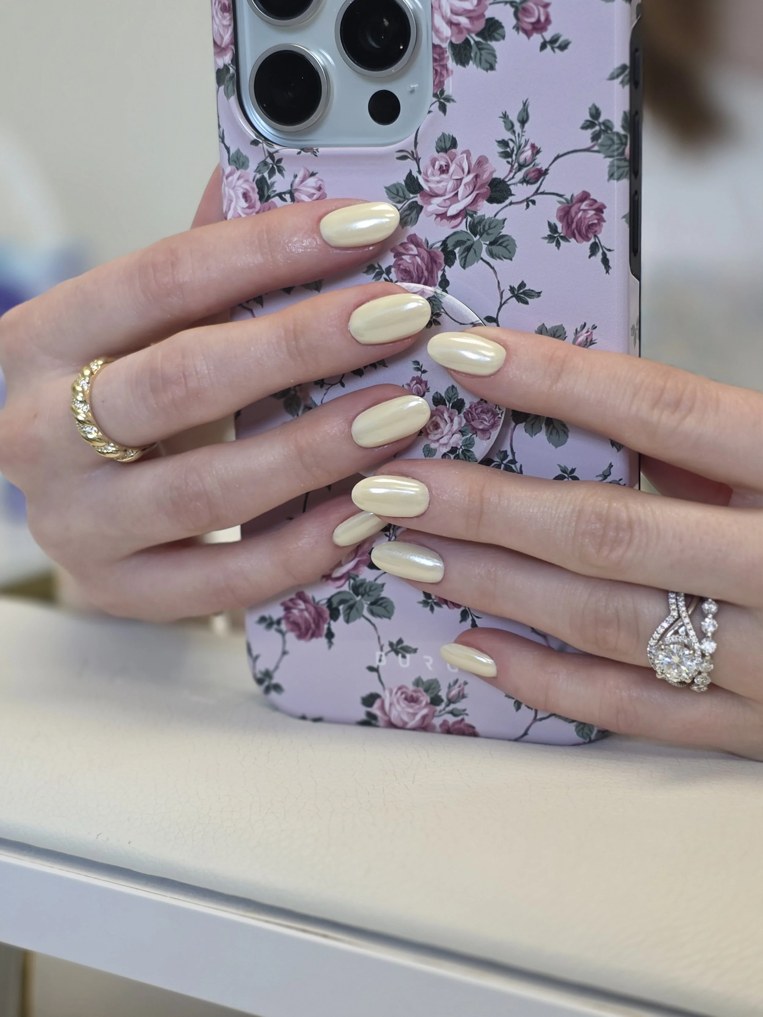 Close-up of a person's left hand with pale yellow nail polish, wearing rings including a gold ring with small diamonds and a diamond engagement ring, holding a cellphone with a floral case featuring pink roses and green leaves, in front of a pink flo