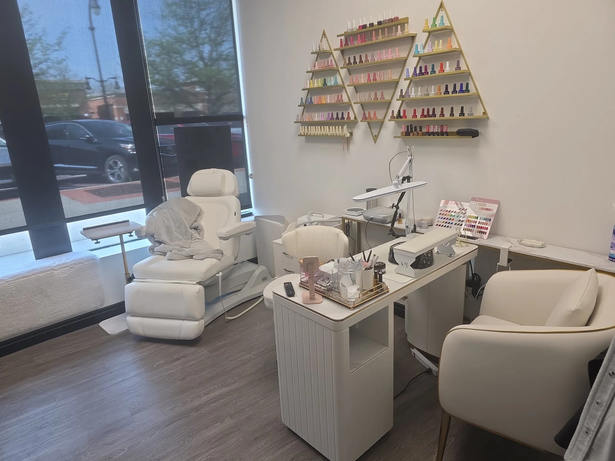 Interior of a modern nail salon with bottles of nail polish on decorative wall shelves, a manicure station with tools, and a pedicure chair near large windows.