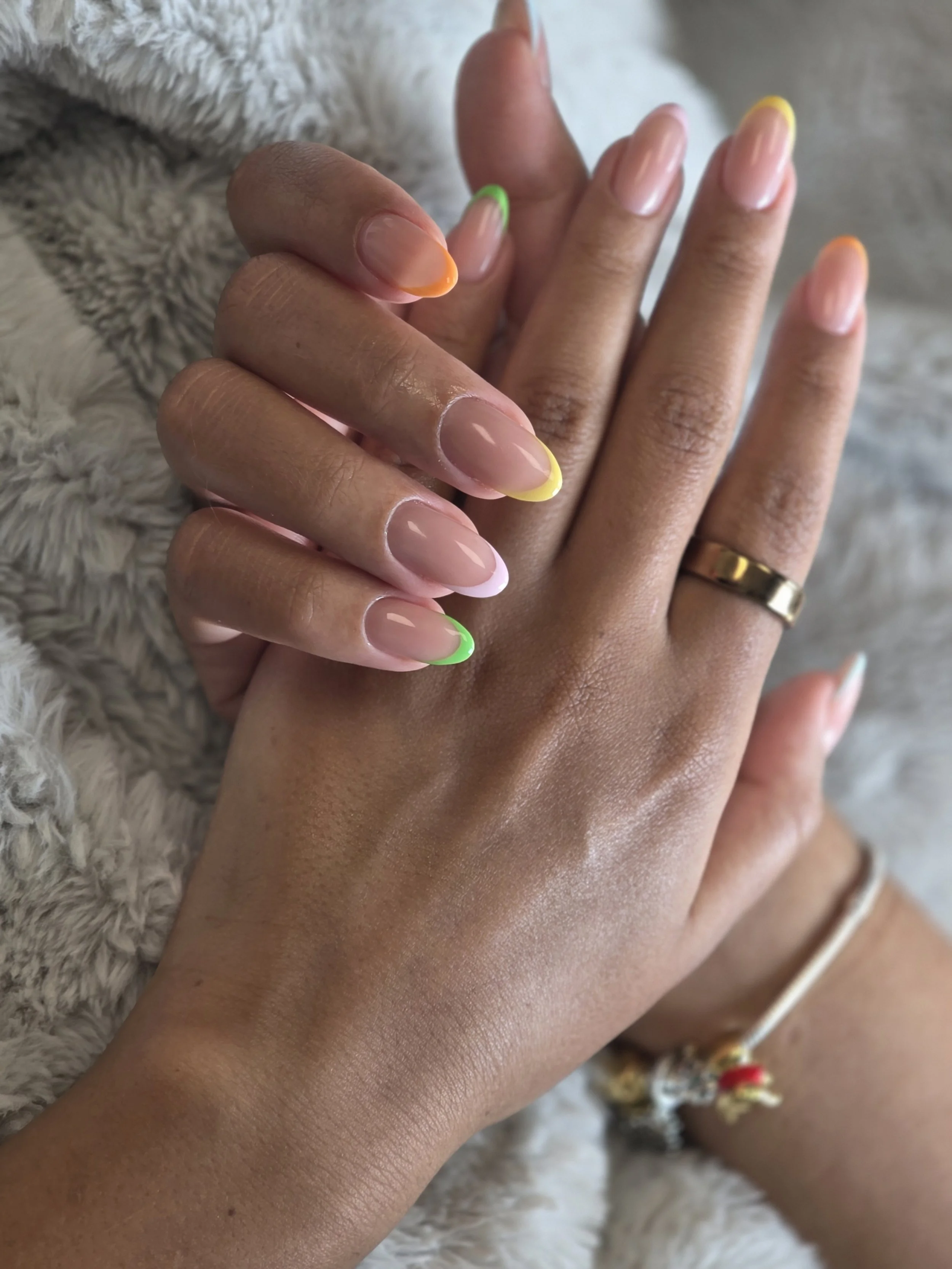 Close-up of two hands with manicured nails in different neon colors, resting on a soft, furry surface, with a ring on one finger.