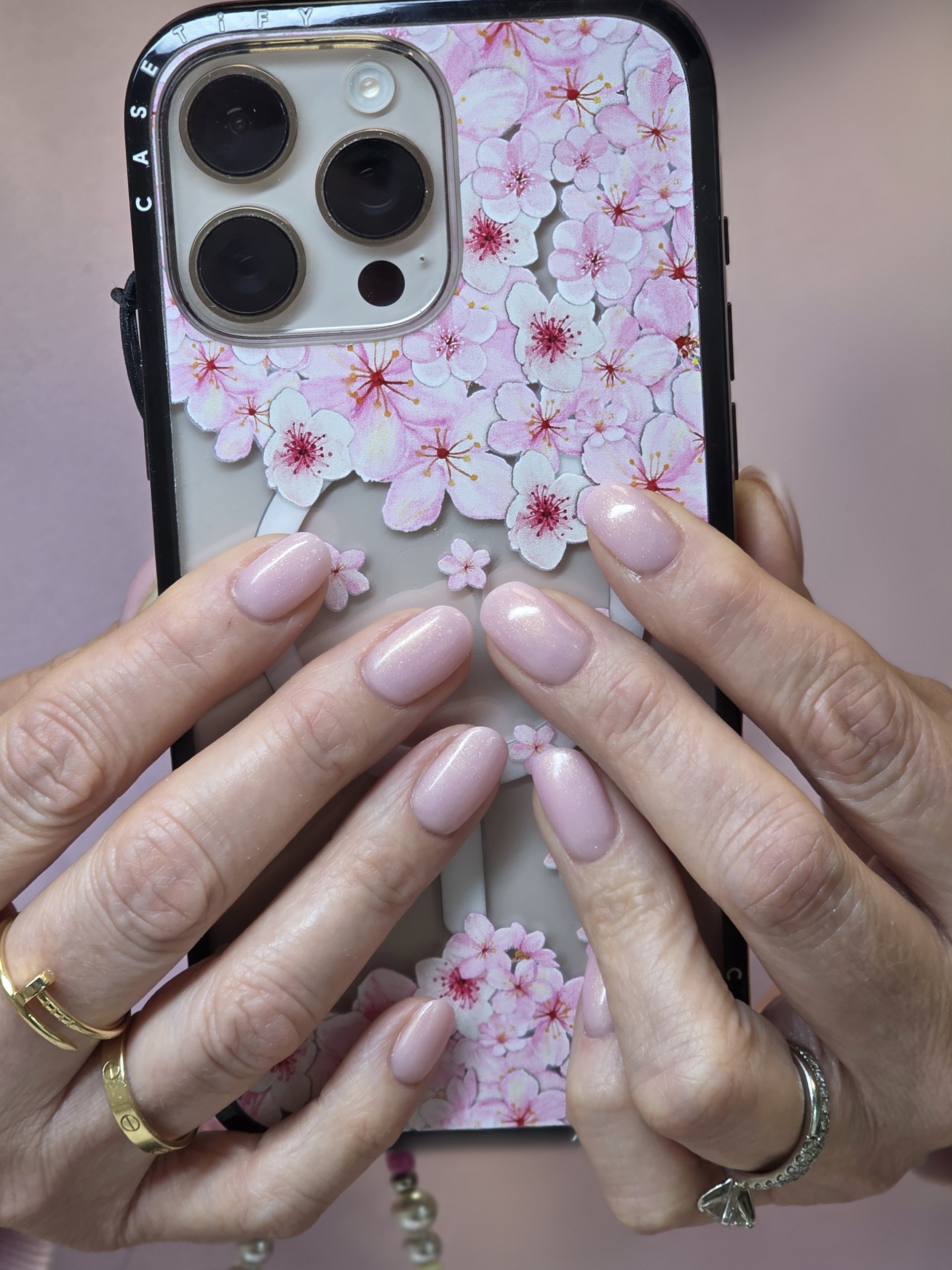 Close-up of hands holding a smartphone with a cherry blossom floral phone case, multiple rings on fingers, with a pearl bracelet, pink nails with glitter.