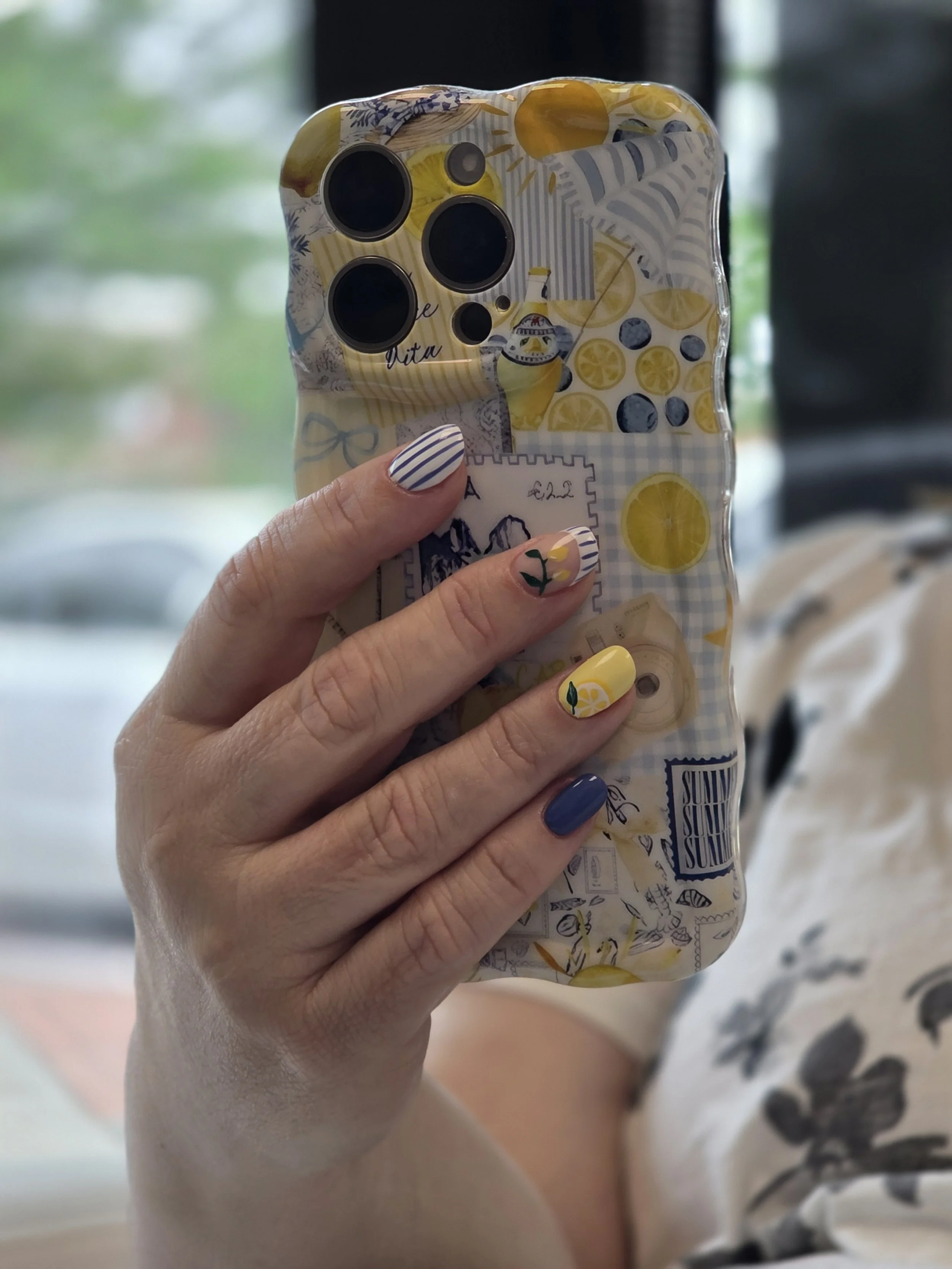 A person's hand with floral painted nails holding a smartphone in a decorative lemon-themed case.