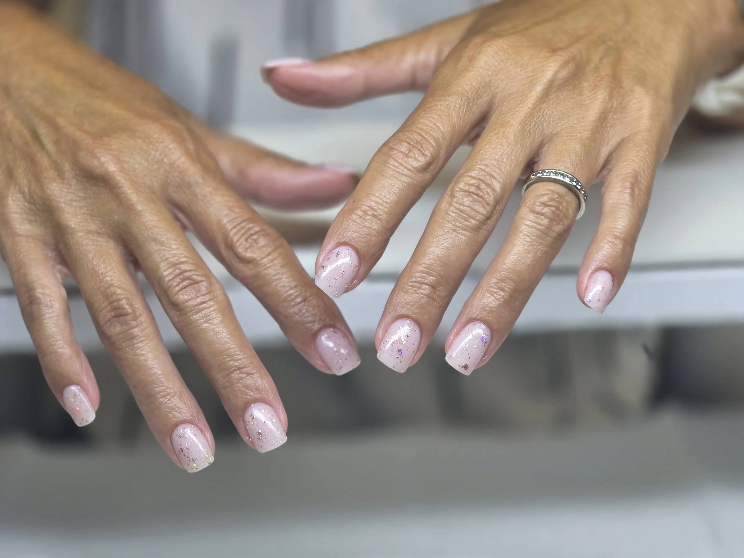 Close-up of a person's hands with decorated nails and a silver ring on the middle finger of the right hand. The nails are painted with light pink polish and sprinkled with glitter.