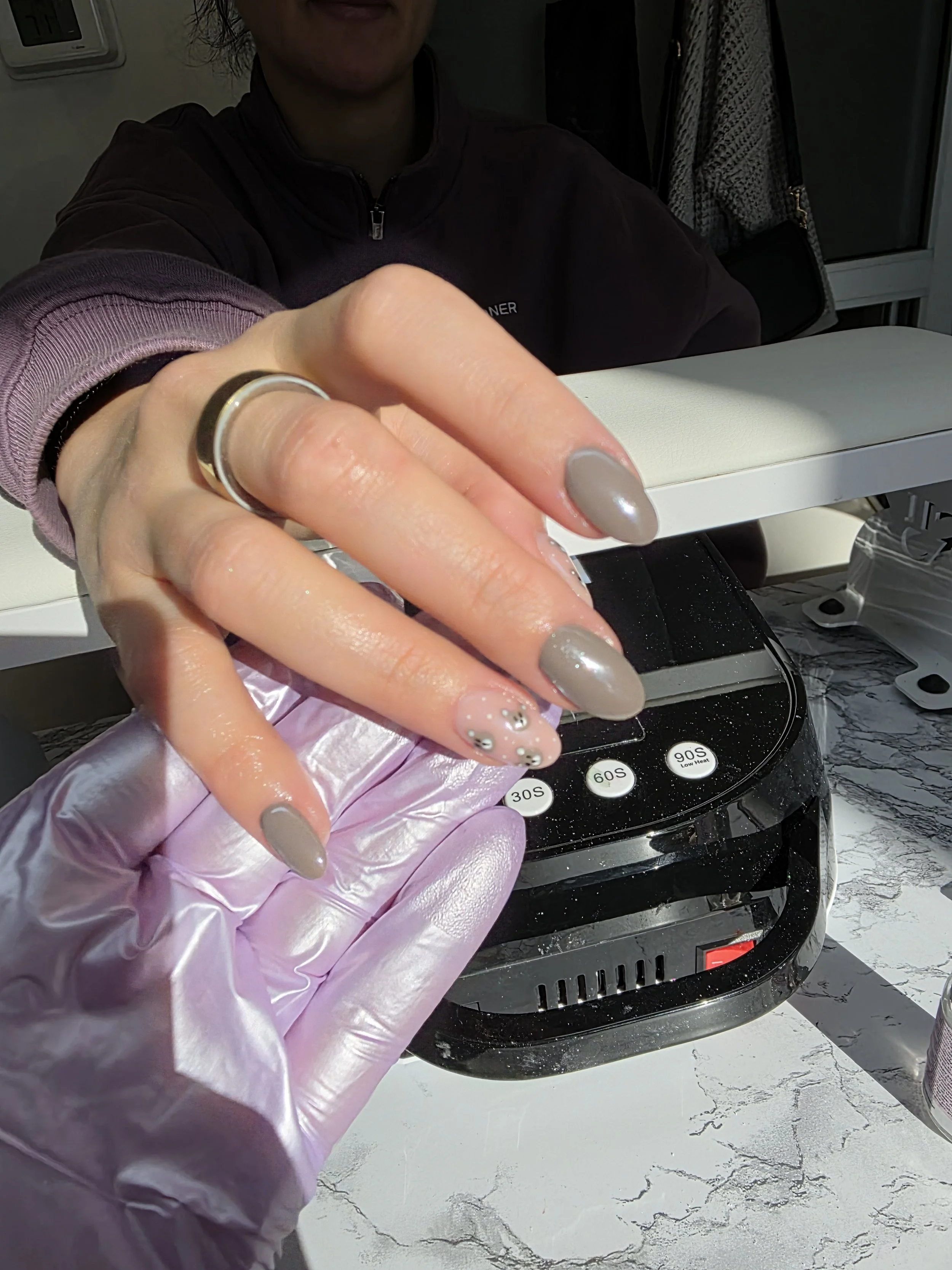 Person with long dark hair wearing a purple hoodie and disposable glove, pointing a smartphone at the camera, with nail art featuring small floral designs, in front of an air fryer oven on a marble countertop.