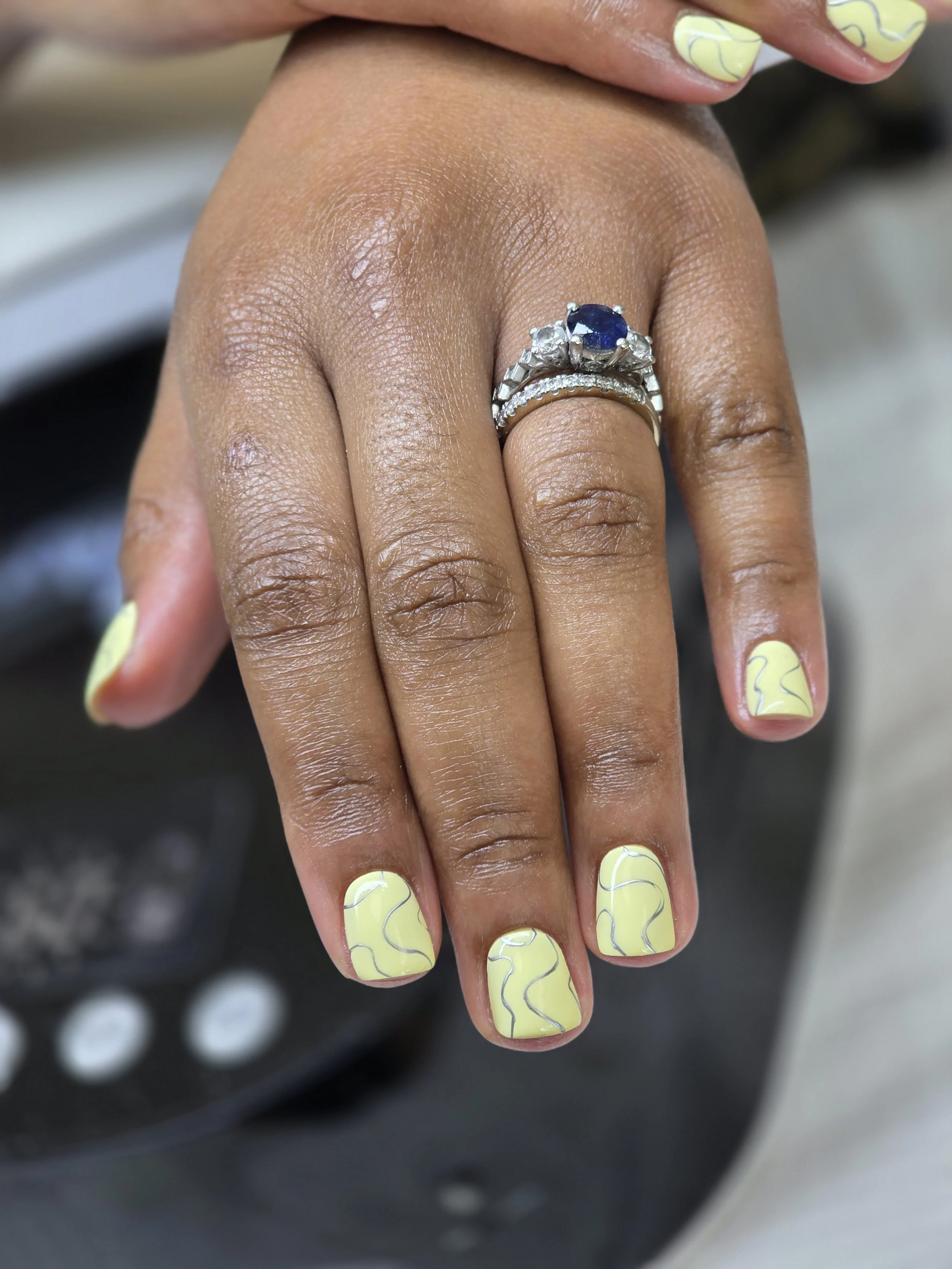 Close-up of a hand with light yellow nail polish featuring black swirl designs. The person is wearing multiple rings, including a silver band with a large blue gemstone and smaller clear stones.