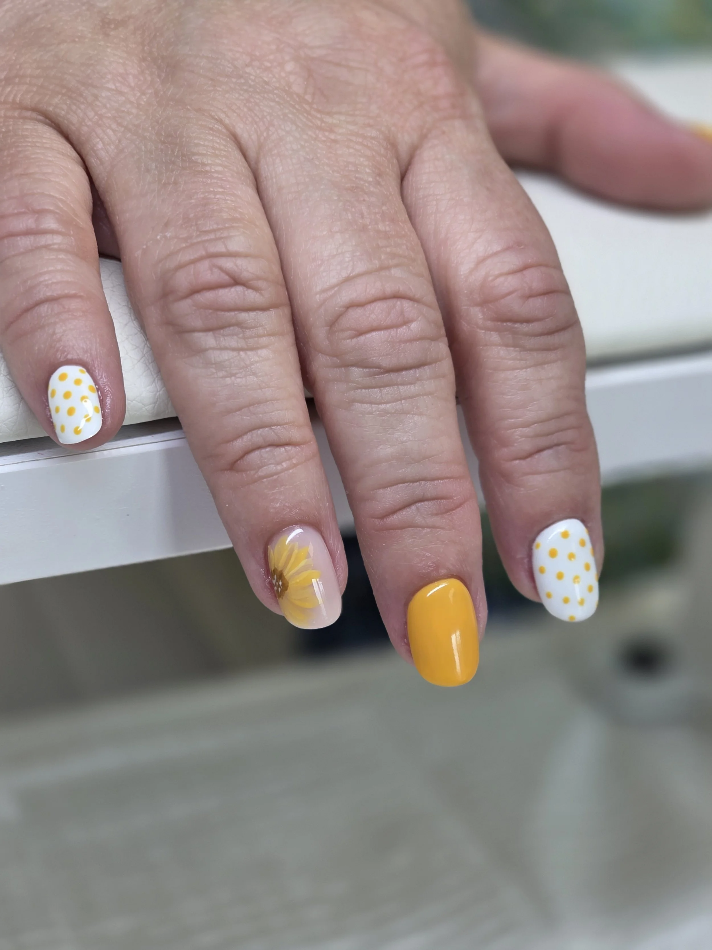 Close-up of a person's hand with yellow and white polka dot nail polish, showing four fingers resting on a white surface.