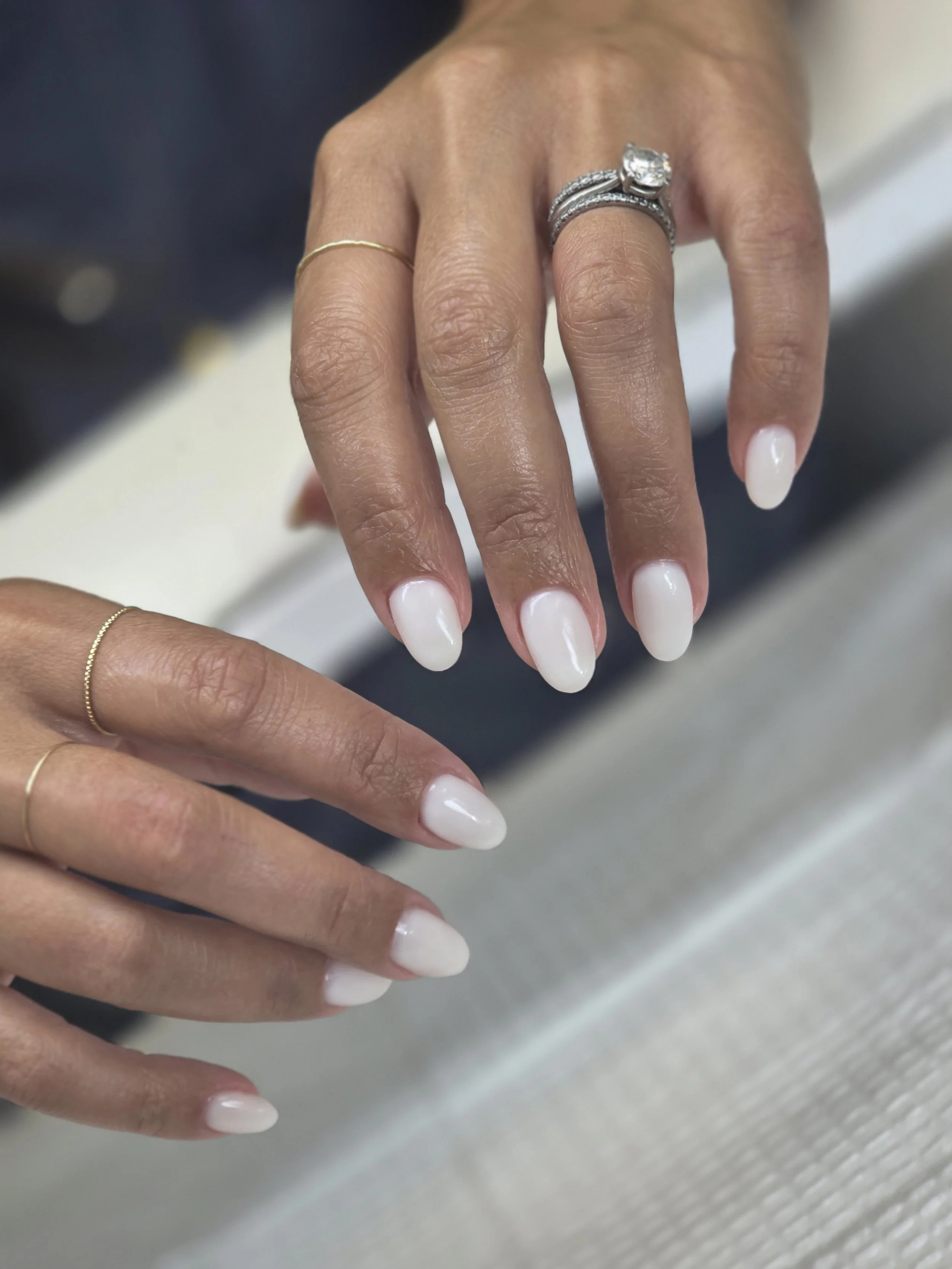 Close-up of a person's hand with white painted nails, several rings including a silver engagement ring with a large diamond, and a thin gold band. The background is blurred.