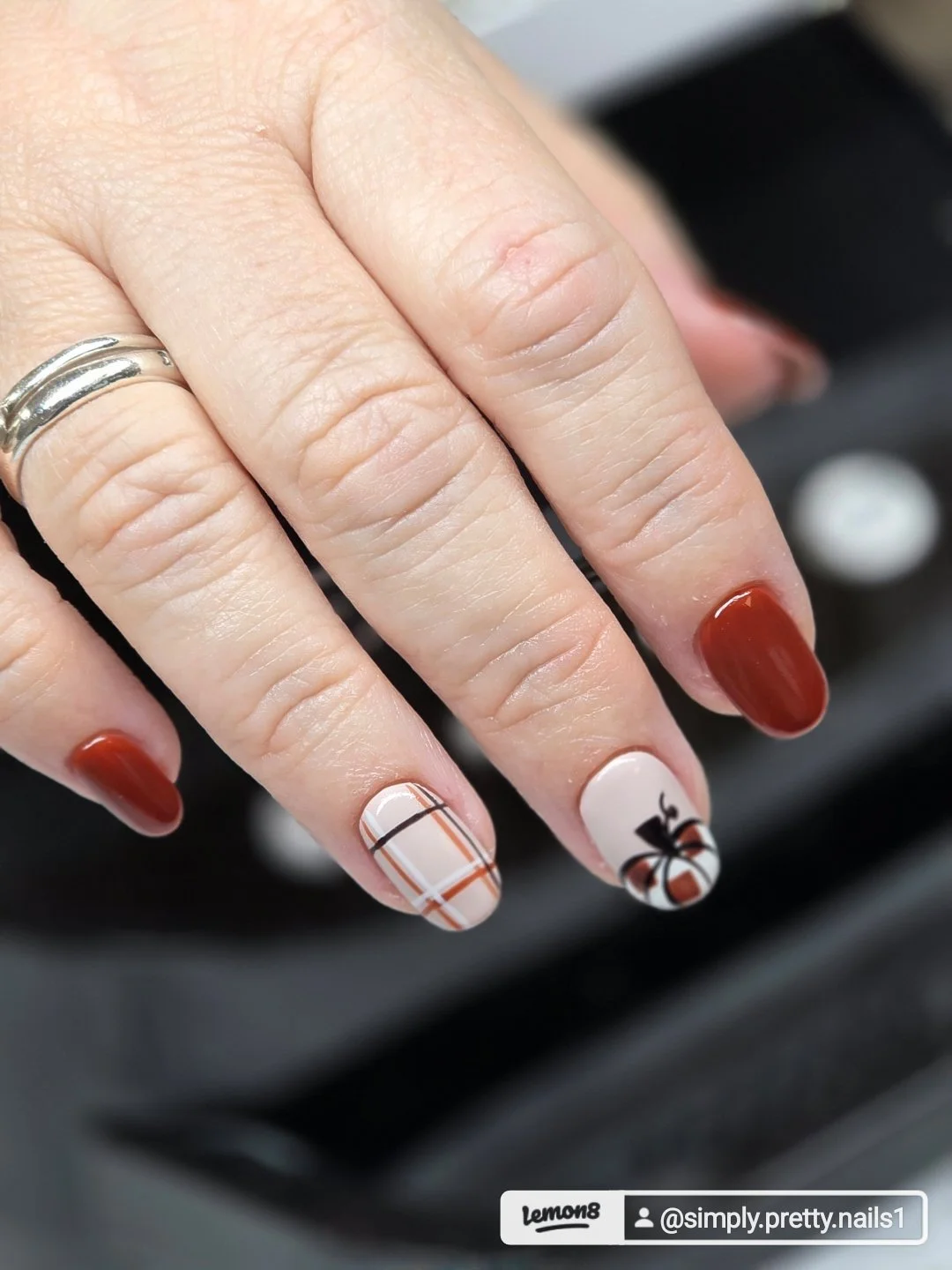 A hand with painted nails and a silver ring, showcasing holiday-themed nail art with red, beige, and plaid patterns.