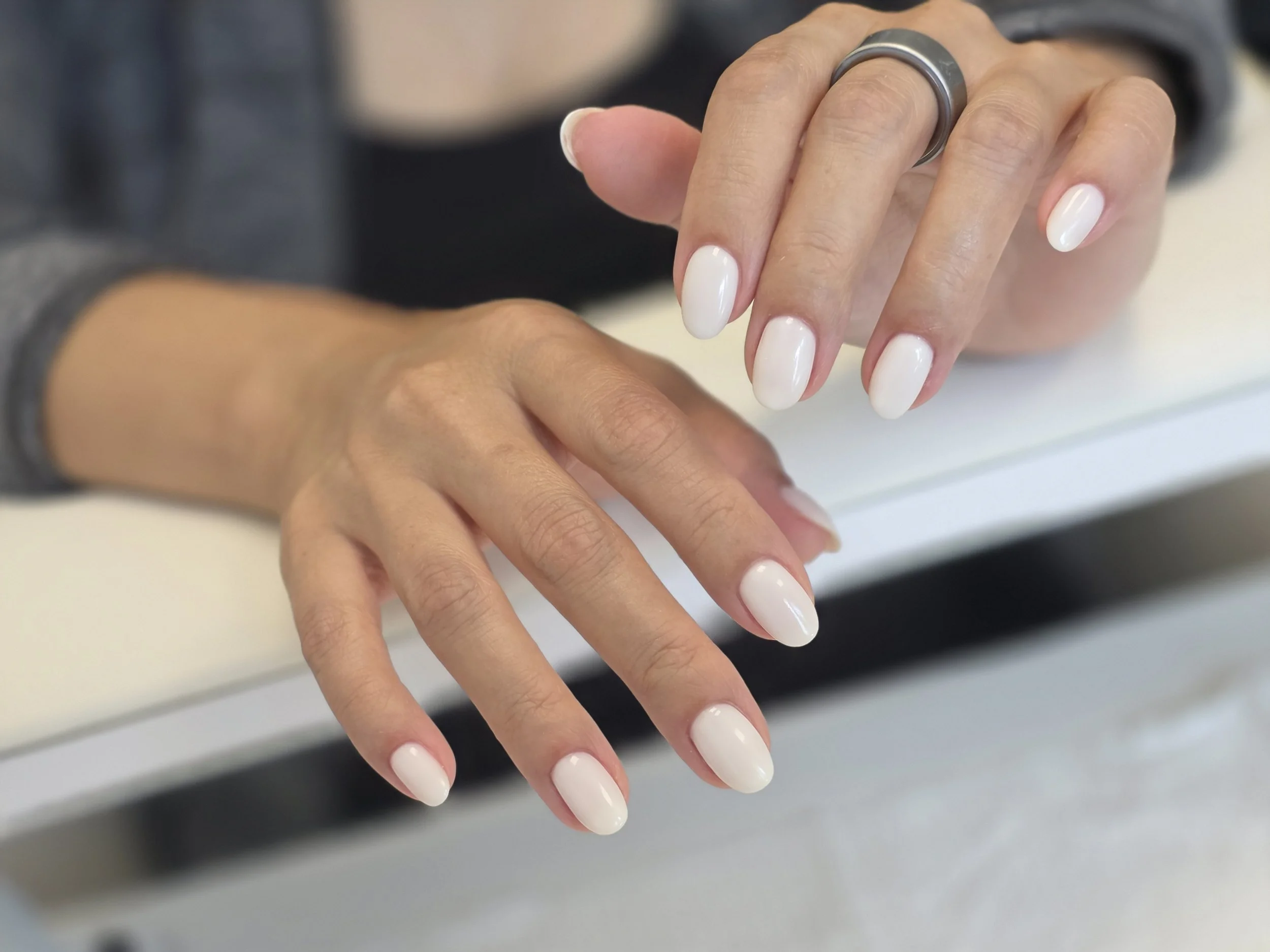 Close-up of a woman's hands with white manicured nails, wearing a silver ring, resting on a white surface.