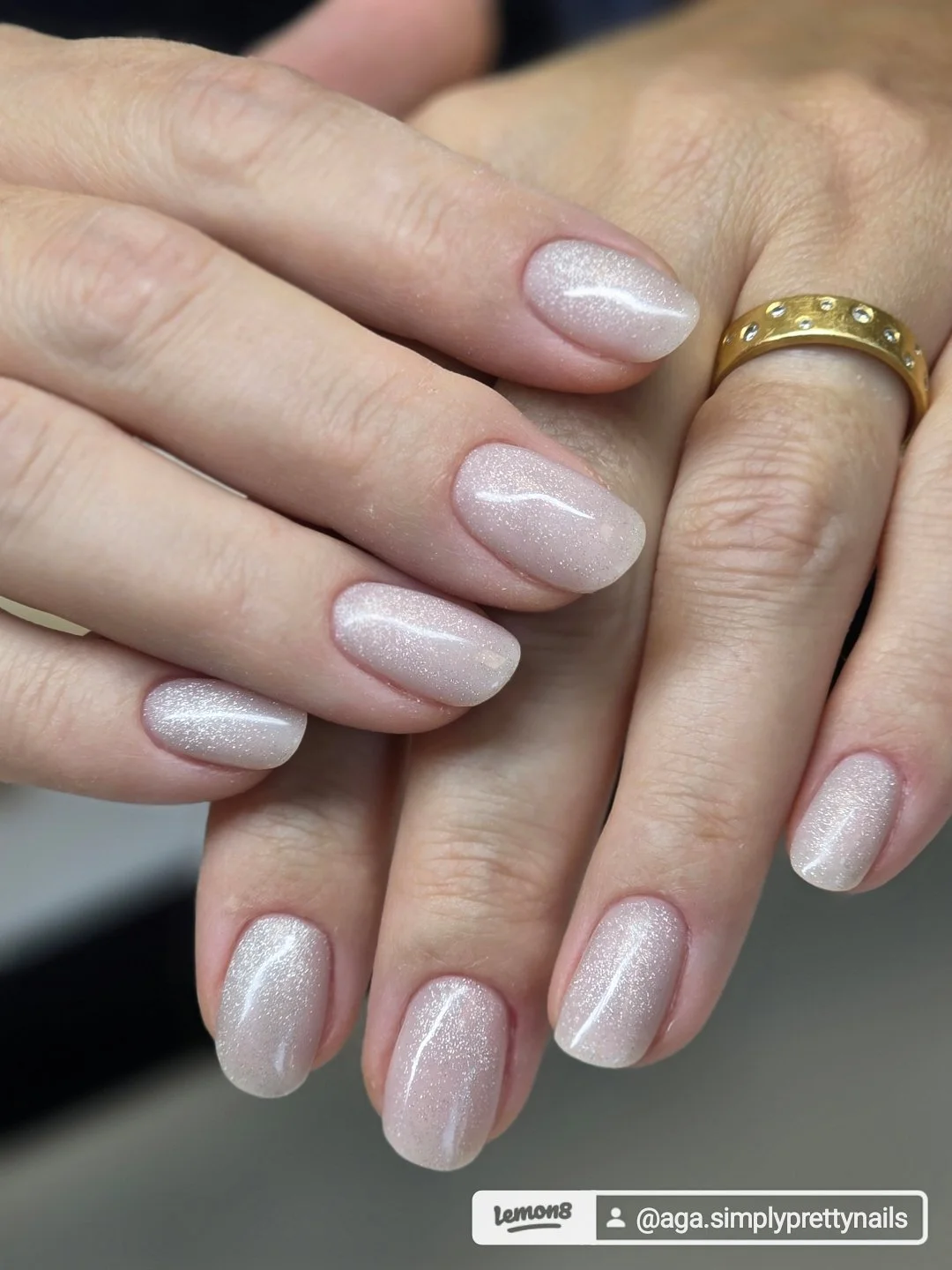 Close-up of hands with manicured nails featuring pale pink, shimmery, glittery nail polish, and a gold ring with small diamonds on the ring finger of one hand.