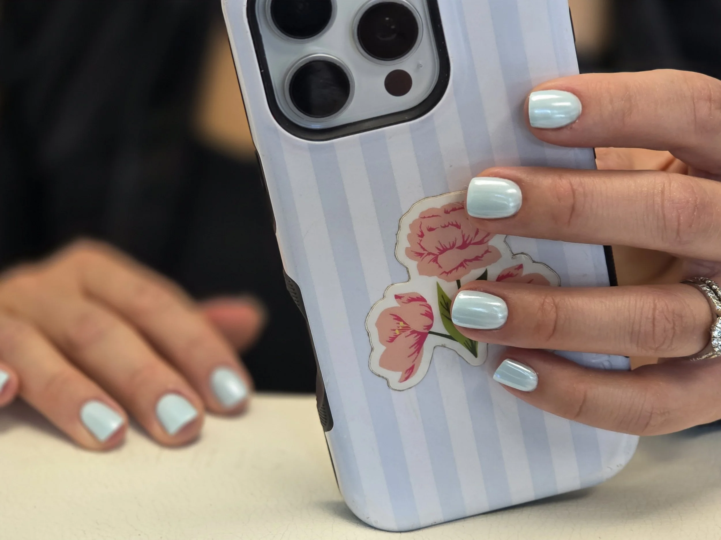 Close-up of a person's hand with light blue nail polish holding a smartphone with a striped case decorated with floral stickers. The background is blurred.