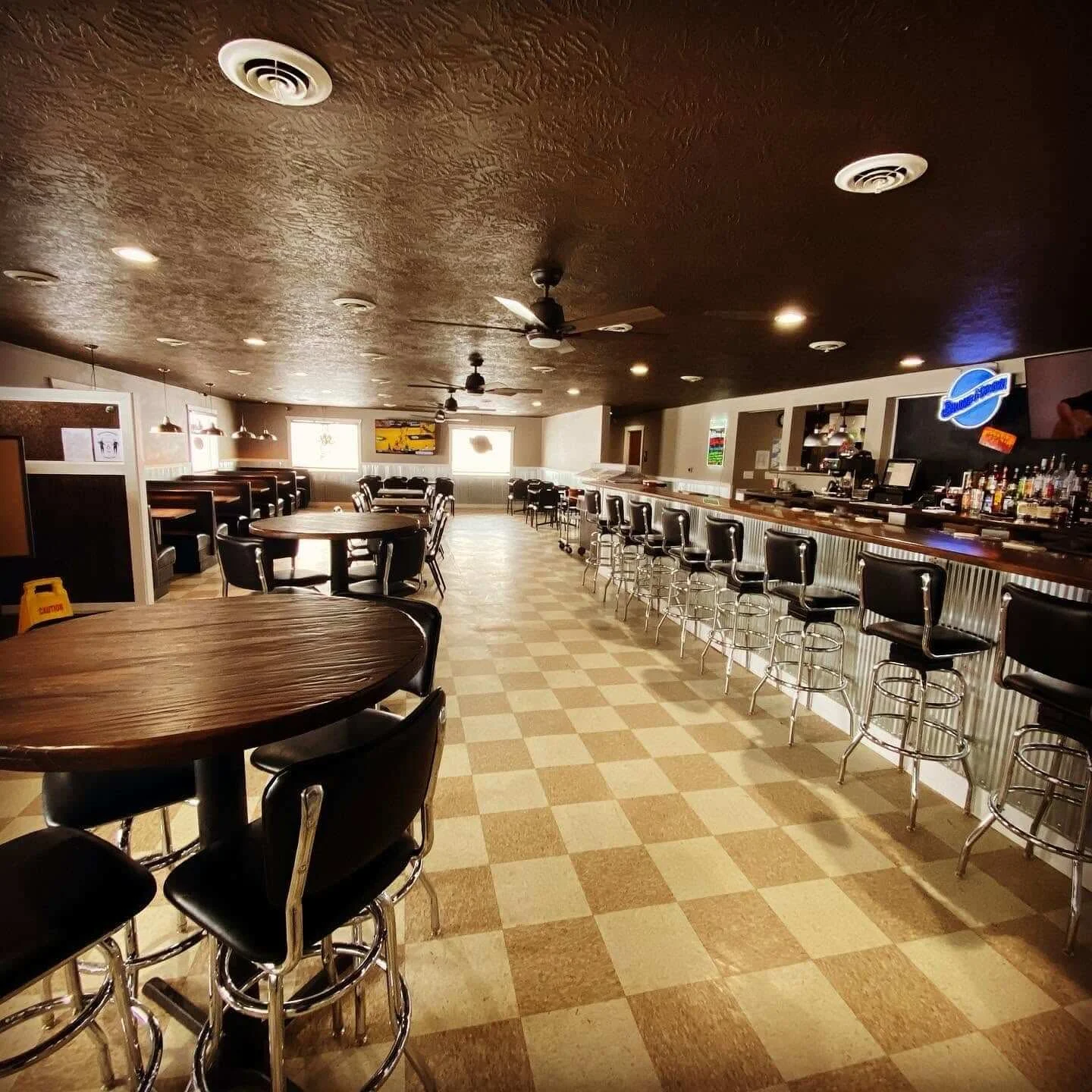 Empty bar and dining area with black chairs, bar stools, and pendant lighting, featuring a bar counter with liquor bottles and a TV in a casual restaurant Buffalo Joe's Roadhouse.