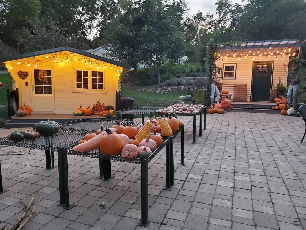 Outdoor patio decorated with string lights, pumpkins, gourds, and gourds arranged on tables and in front of small buildings, celebrating fall or Halloween.