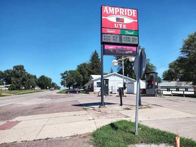 A roadside sign displaying fuel prices and a pizza shop advertisement in a small town with trees, buildings, and a clear blue sky.