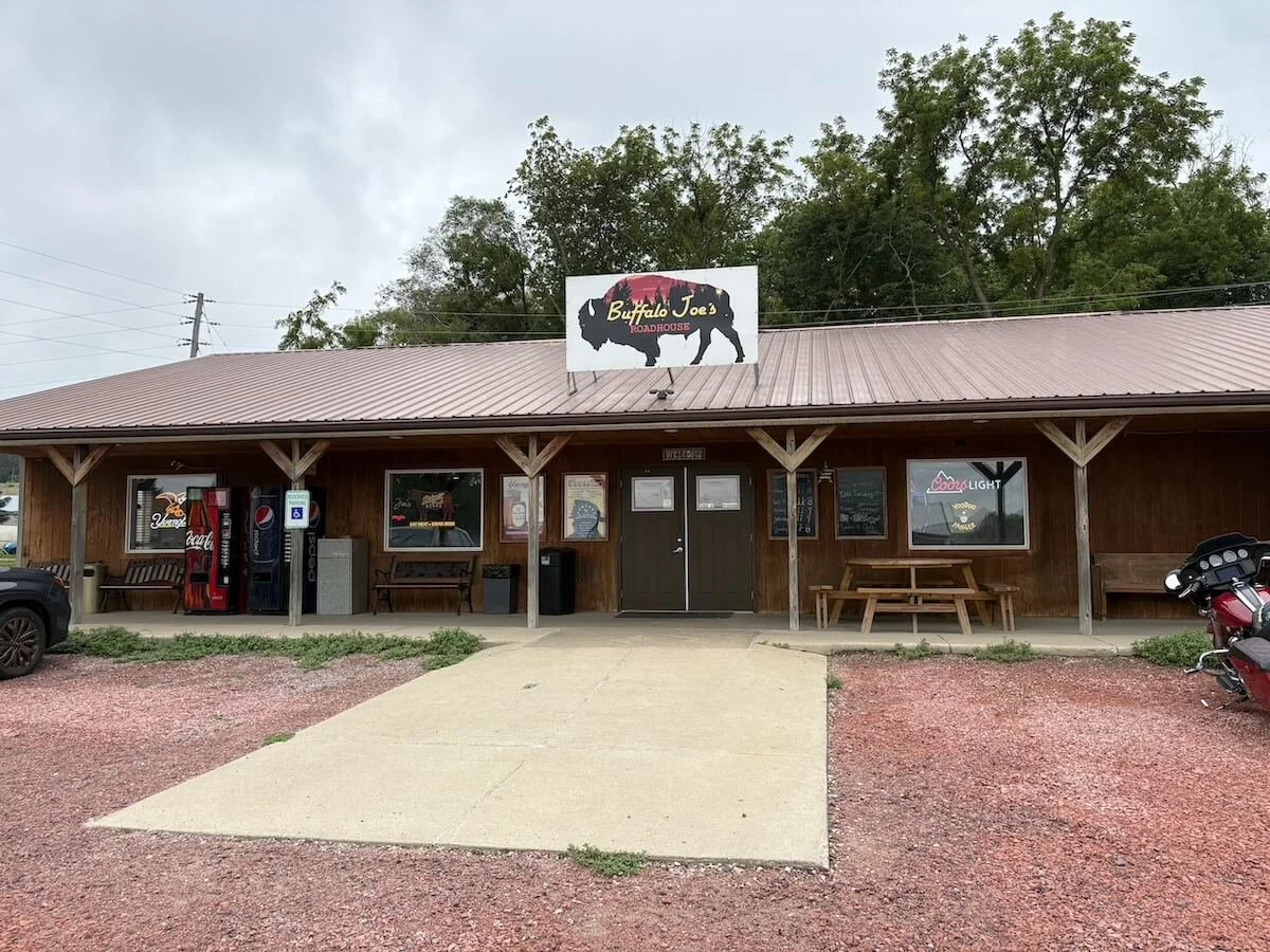 Front view of Buffalo Joe's Roadhouse, a rustic building with a metal roof, wooden exterior, neon signs, vending machines, picnic tables outside, and a motorcycle parked to the right.