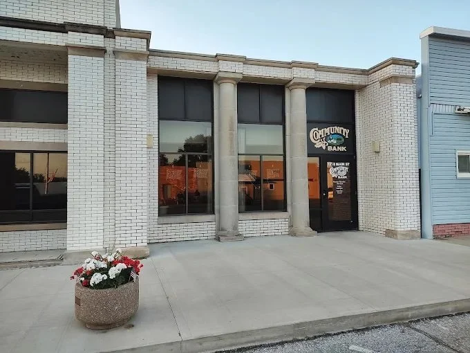 Exterior of a building with large windows, white brick walls, and two columns, with a flower pot filled with white and red flowers in front, and a sign for Community Bank.