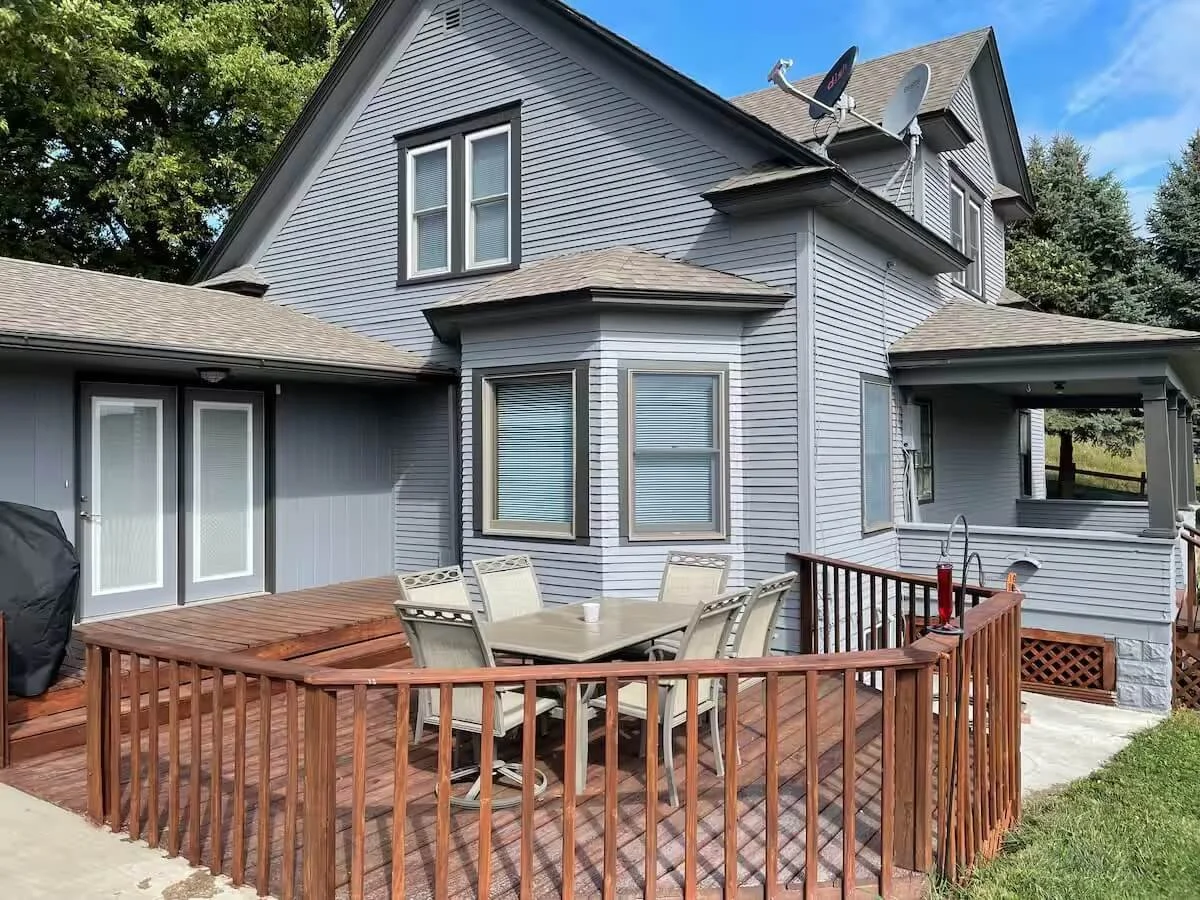 Backyard deck with table and chairs, attached to a gray house with multiple windows and satellite dishes on the roof.
