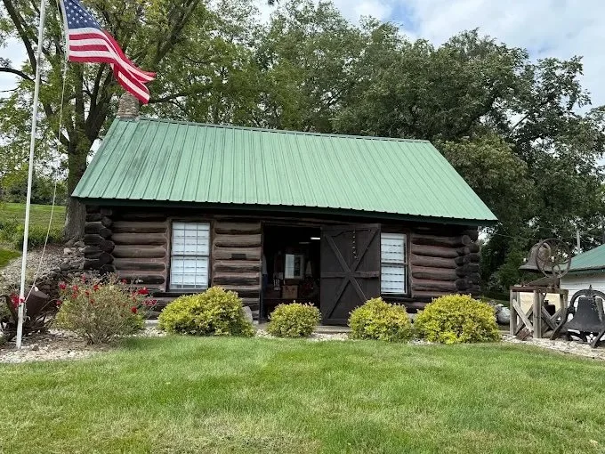 A small log cabin with a green metal roof, black door, and white-framed windows. An American flag on a flagpole is to the left, with well-manicured bushes and a lawn in front. There are trees and a cloudy sky in the background.