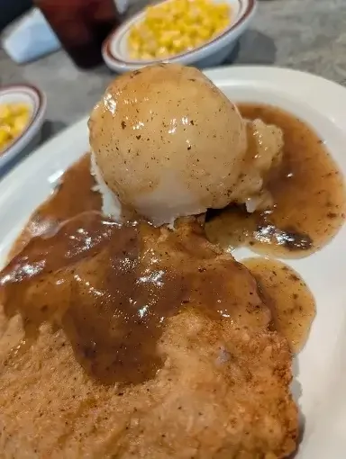 Assorted food on a white plate including a piece of fried chicken with gravy, a scoop of mashed potatoes with gravy, and a dish of corn on the cob in the background.