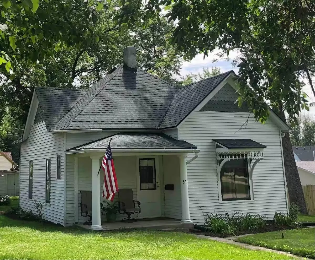 A small white house with a front porch, American flag, and flower bed in the yard, surrounded by trees.
