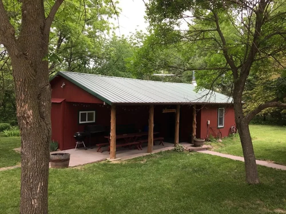 A red wooden building with a metal roof, surrounded by green trees and grass, with a covered porch area that has picnic tables and a small round grill.