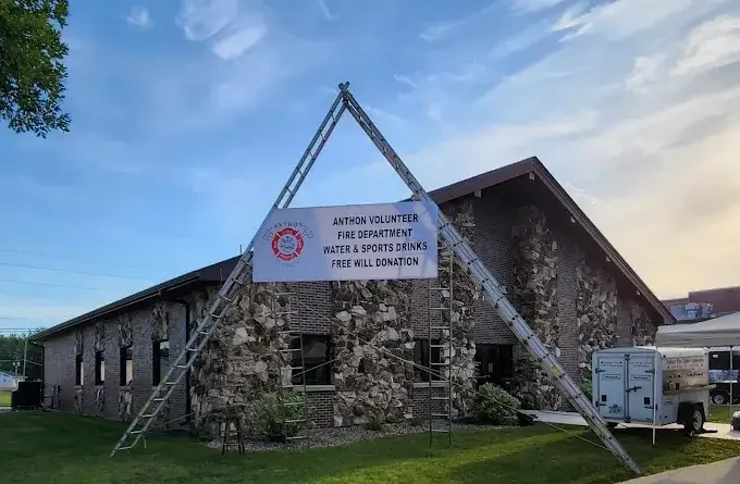 A large building made of stones with a sign hanging from two ladders reading "Anthony Volunteer Fire Department Water & Sports Drinks Free Will Donation." The scene is outdoors with a grassy area and a trailer nearby.