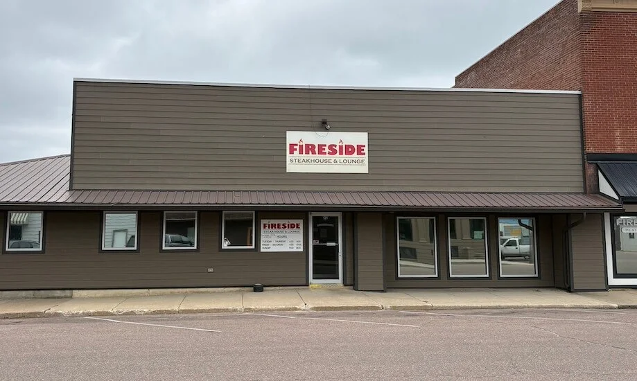 The exterior of a restaurant called Fireside Steakhouse & Lounge, with a large sign on the building's front and smaller sign next to the door displaying opening hours.