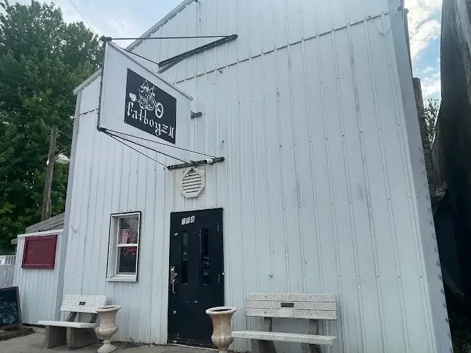 White building with a black sign that says 'Peter A. Hansen Post *365' and a memorial front righ with an American flag and two small statues honoring veterans. The building has a black door, a window, two benches, and two stone planters outside.