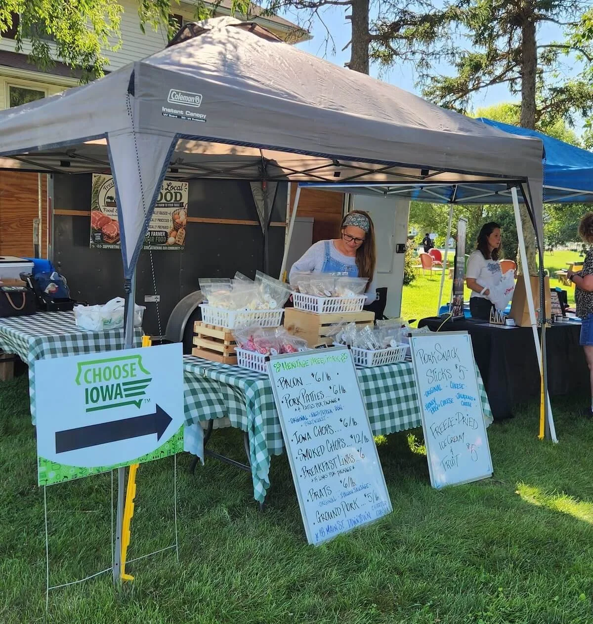 A farmers market stall set up under a gray canopy with a green and white checkered tablecloth, selling pork products. Two women are attending the stall, one inspecting items and the other holding a bag. Handwritten signs display prices for bacon, pork patties, smoked chops, breakfast links, and other smoked meats. A sign with an arrow and 'Choose Iowa' directs visitors. The market is outdoors on a grassy area with trees and houses in the background.