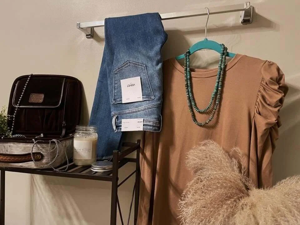 Clothing and accessories setup including pair of blue jeans hanging on a rack, a beige dress with ruffled sleeves on a blue hanger, a beaded necklace, a black handbag, a candle, a small plant, and a decorative beige pampas grass arrangement.