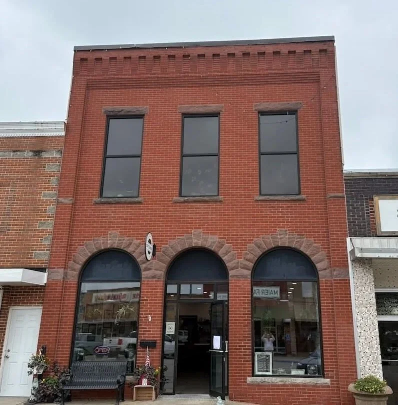 A three-story red brick building with arched windows on the ground floor and rectangular windows on the second floor. There is a storefront entrance with glass doors in the center and a black bench outside. The building is situated between other brick buildings.