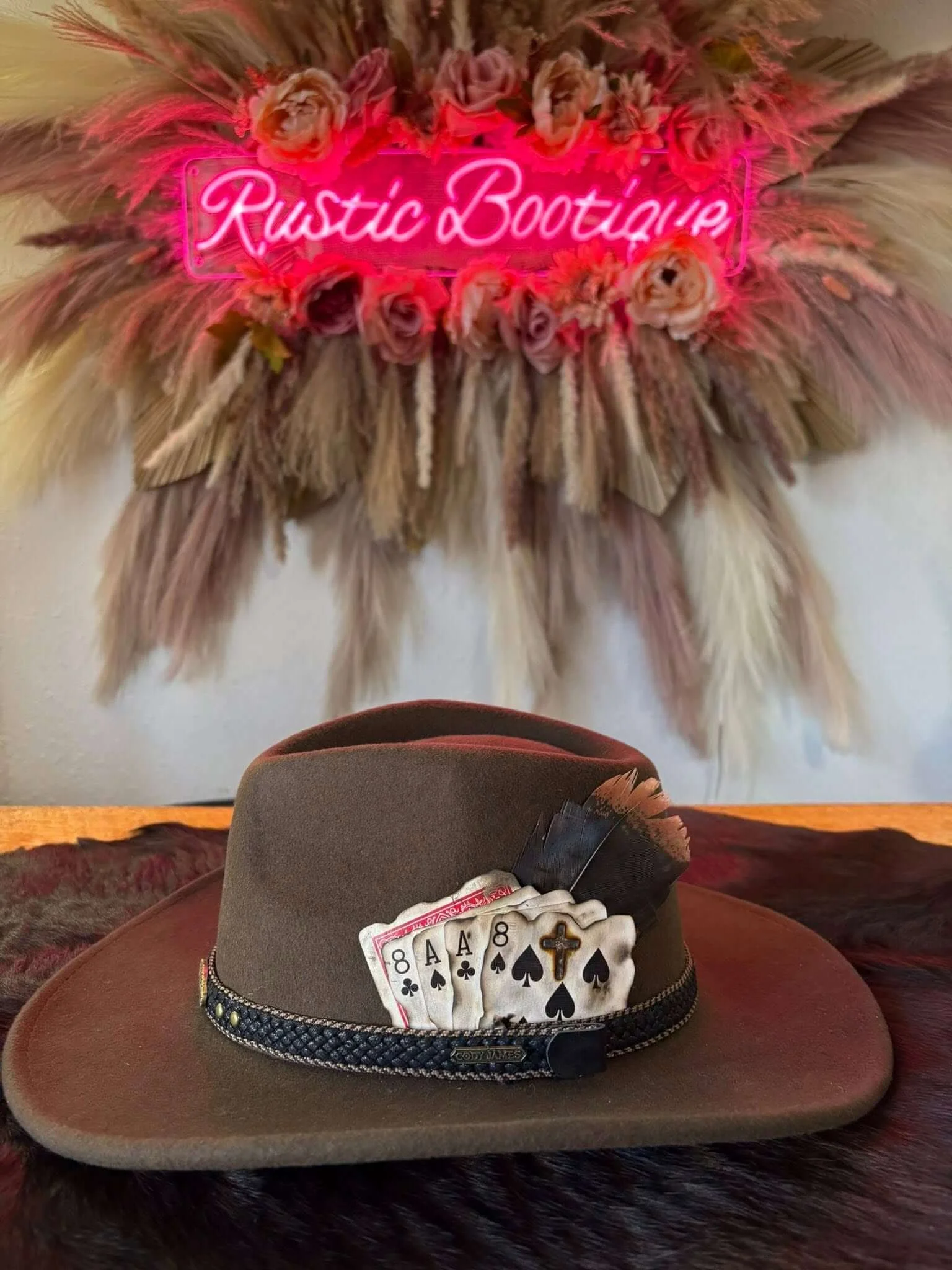 A cowboy hat with playing cards, a cross, and a bandana, placed on a felt surface with a decorative feather, in front of a large feathered wall hanging with a neon sign that reads 'Rustic Boutique' surrounded by pink and beige roses.