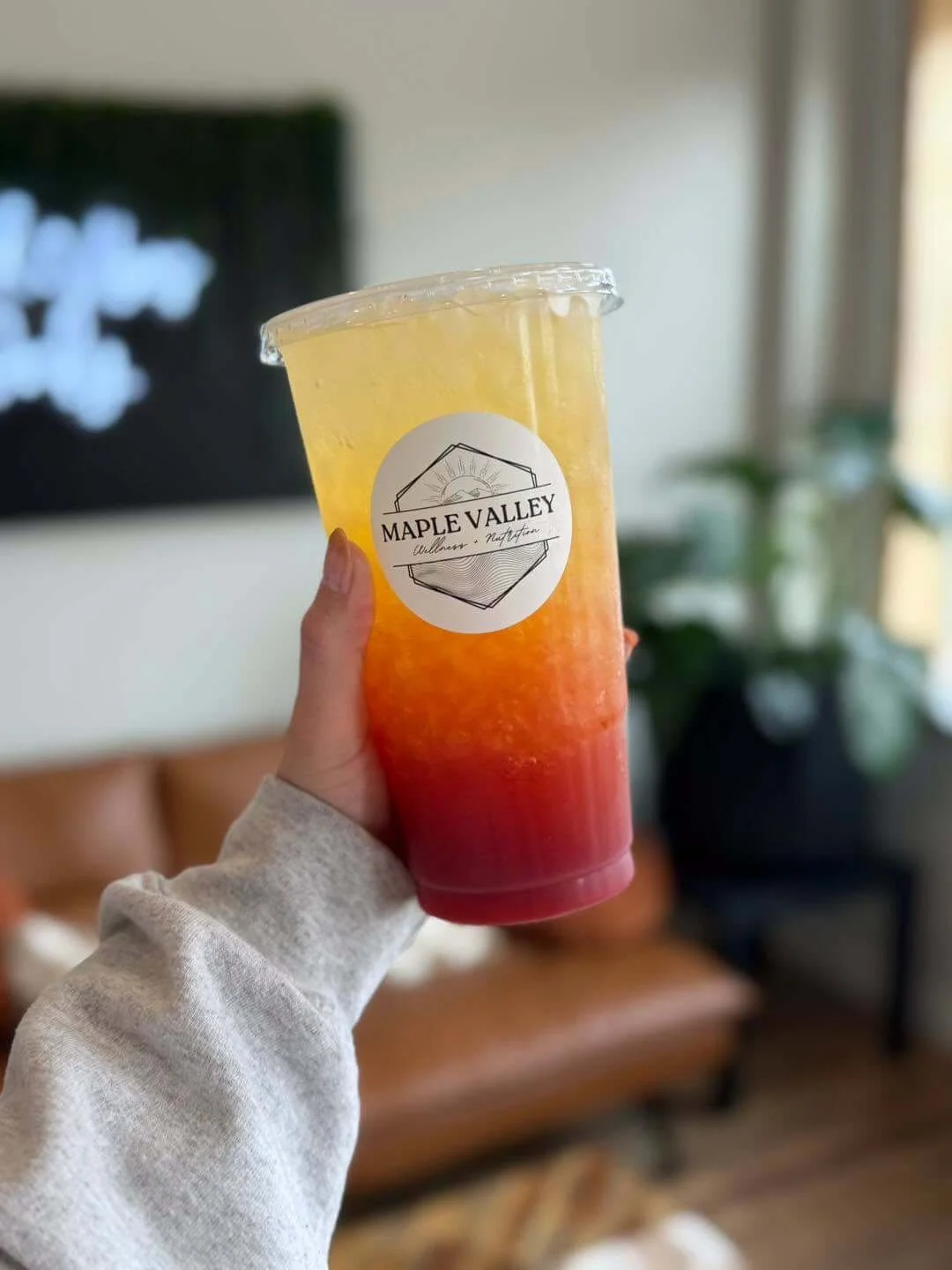 A hand holding a colorful layered drink with red, orange, yellow, and clear sections in a plastic cup with a Maple Valley logo, in a cozy indoor setting.