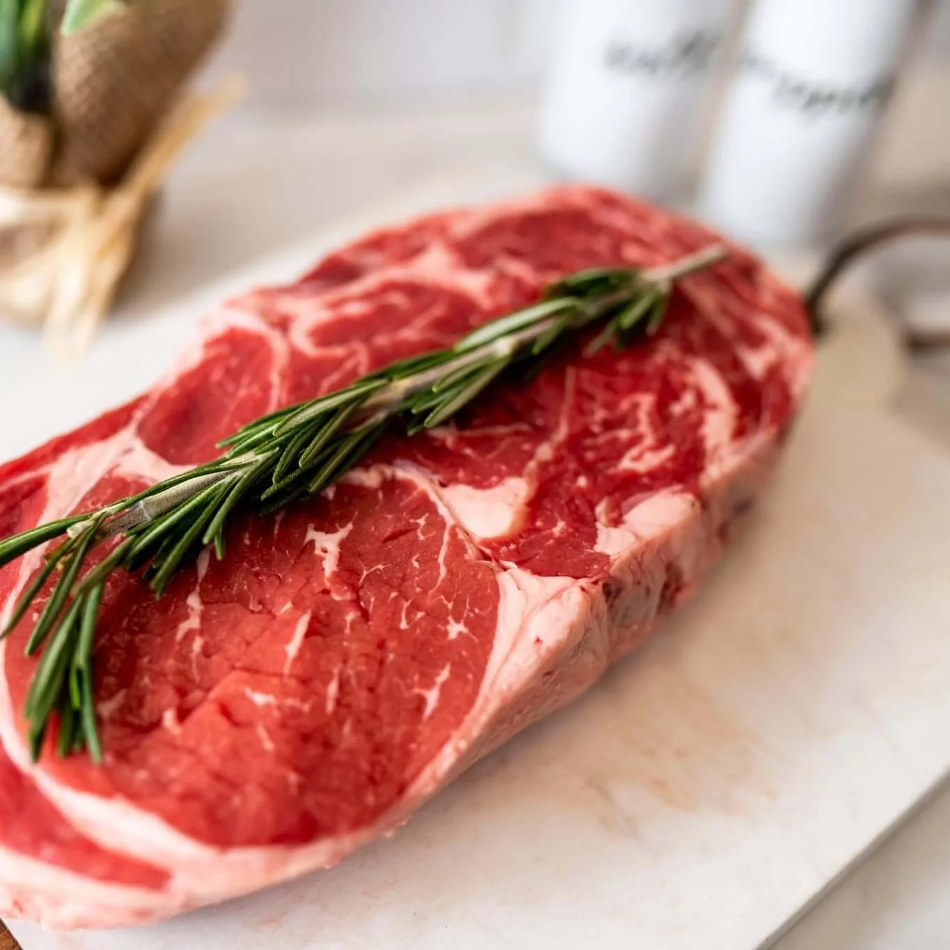Raw beef steak with a sprig of fresh rosemary on top, placed on a white surface.