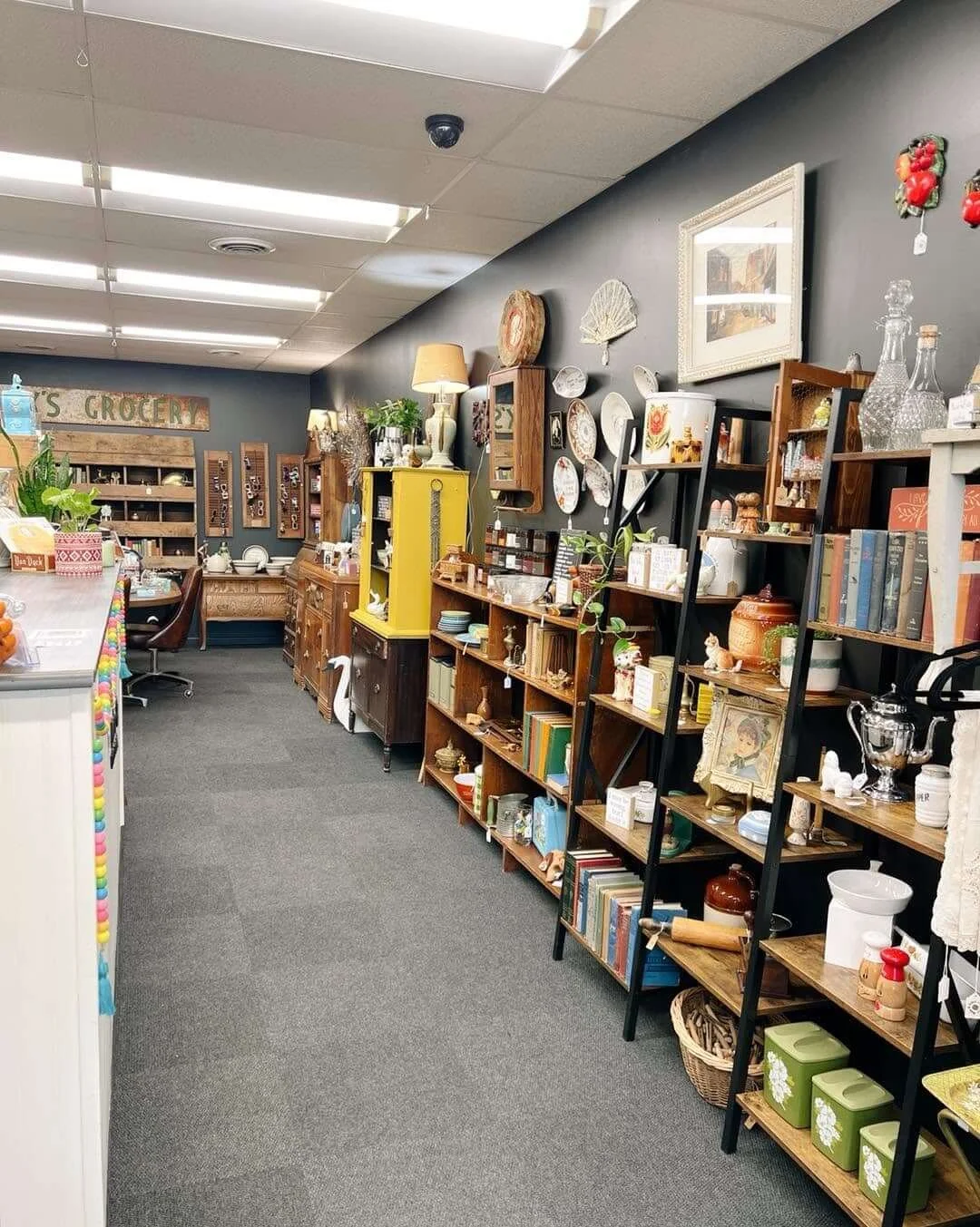 Side aisle of a thrift store with wooden shelves filled with various vintage and antique items, including books, ceramics, glassware, and decorative pieces, with a dark wall in the background and bright overhead lighting.