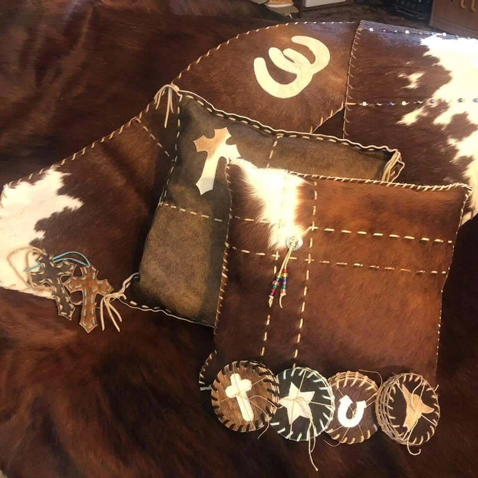 Four square pillows with cowhide patterns and stitched borders, decorated with Christmas-themed ornaments such as wooden crosses, stars, and letters spelling 'Jesus', placed on a cowhide surface.