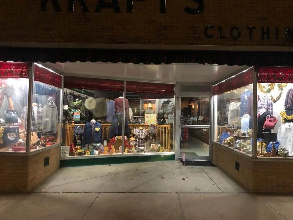 A storefront clothing store at night with large glass windows showcasing mannequins dressed in various outfits and accessories, illuminated inside with yellow and white lighting.