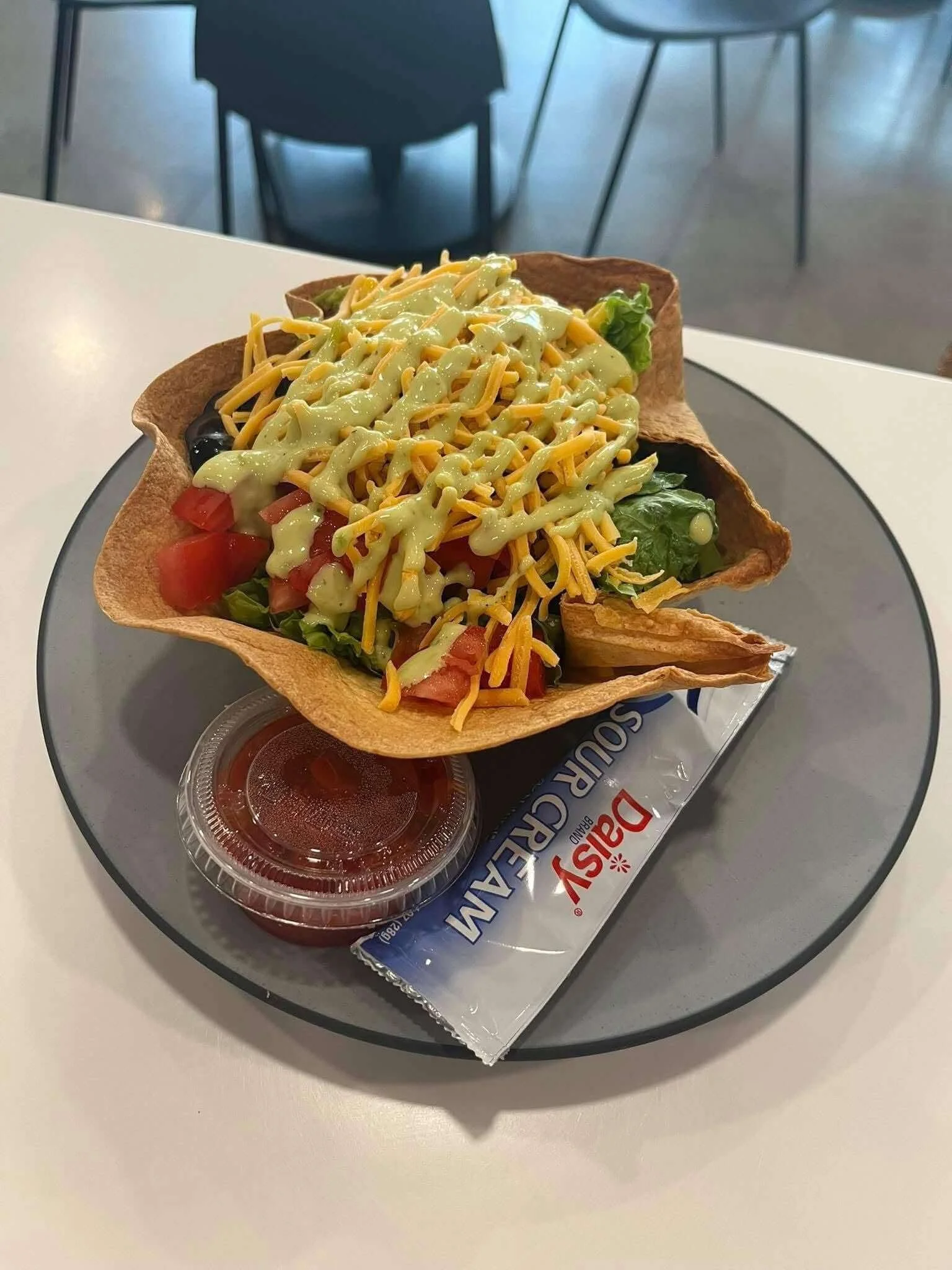 A taco shell filled with lettuce, diced tomatoes, shredded cheese, and topped with ranch dressing, served with a side of salsa in a clear container.