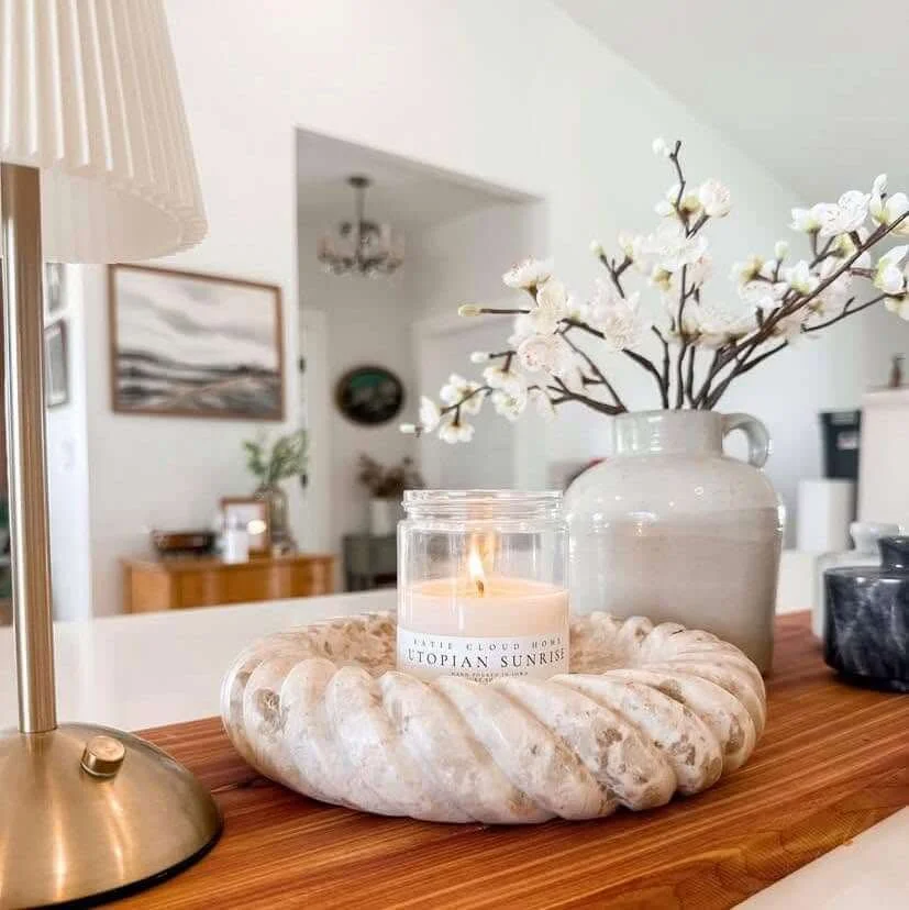 Decorative candle in glass jar on a rock platter with white flowers in a vase on a wooden surface.