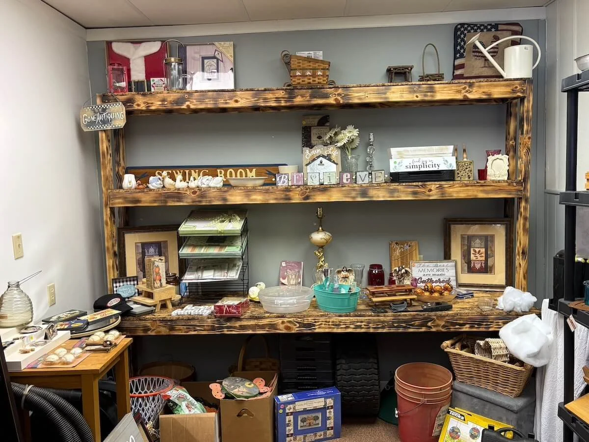 A large wooden shelving unit filled with various decorative items, framed pictures, and small collectibles, with a table underneath holding more items and storage boxes around it.