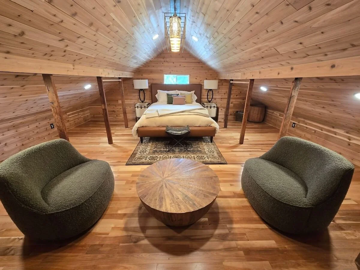 Wood-paneled attic bedroom with a bed, two nightstands with lamps, two green armchairs, a round wooden coffee table, and a small window.