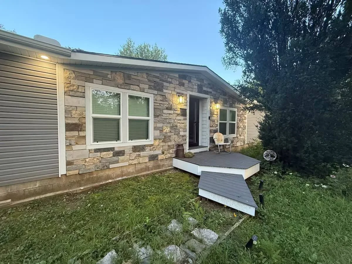 Backyard view of a house with stone exterior, white trim, two windows, a small porch with chairs, and a grassy yard surrounded by trees.