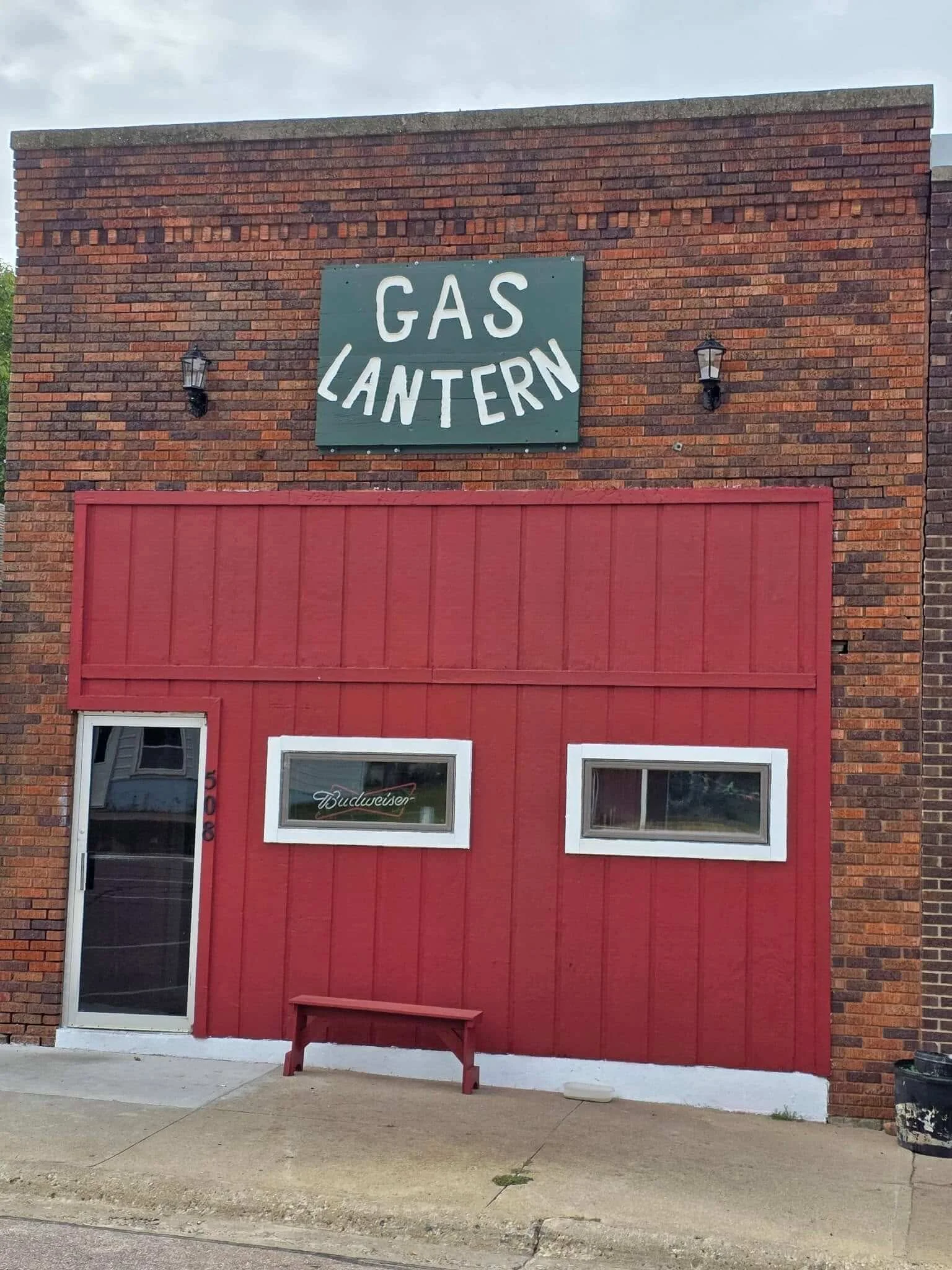 A brick building with a sign reading 'Gas Lantern' on the upper wall, two lantern-style lights on either side of the sign, a red wooden panel below the sign, a glass door with a 'Budweiser' sticker, and a small red bench outside. The sky is cloudy.