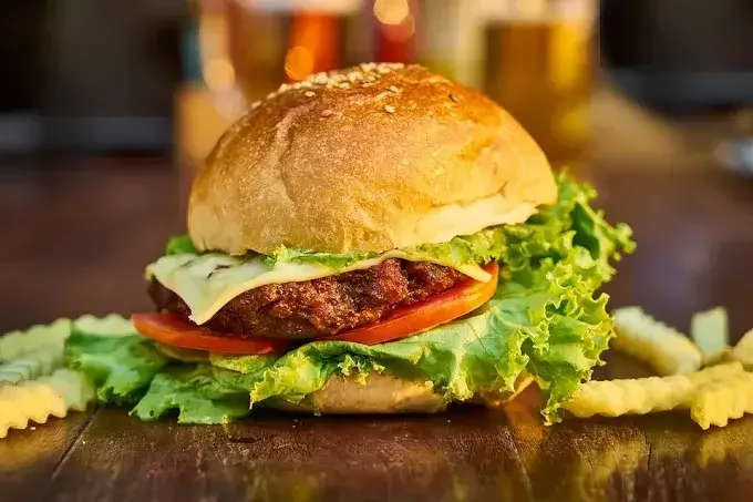 Close-up of a hamburger with lettuce, tomato, cheese, and a beef patty in a bun, with crinkle-cut fries on a wooden surface.