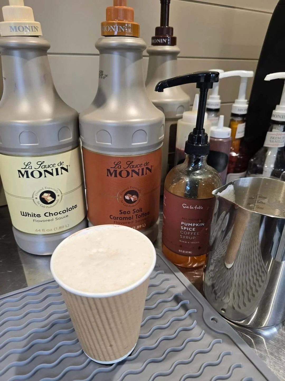 Close-up of coffee syrups, including White Chocolate, Sea Salt Caramel Toffee, and Pumpkin Spice, next to a paper cup filled with frothy coffee, on a gray rubber mat at a coffee shop counter.