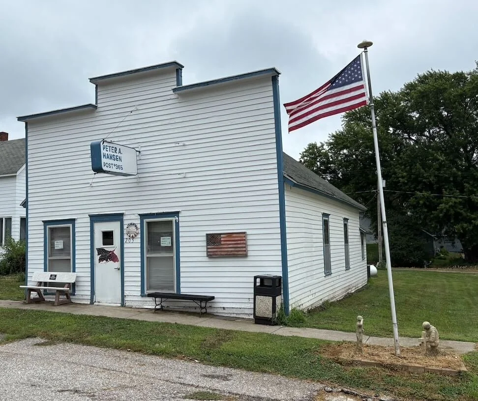 A small white building with blue trim, American flags, benches, a trash bin, and patriotic decorations, with a sign that reads 'Peter A. Hansen Post 365.'