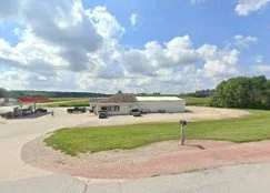 A gravel driveway with a grassy area and a mailbox in a rural setting. Several buildings and vehicles are visible in the background under a partly cloudy sky.