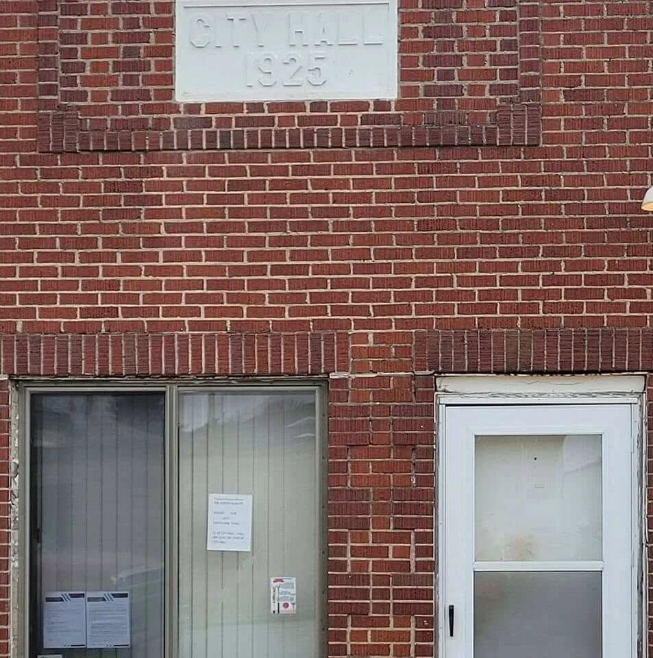 Red brick building with a white sign reading 'City Hall 1925' at the top. Below are a window with vertical blinds and a door with a panel, both on the brick facade.